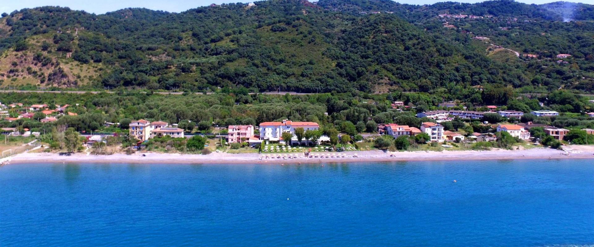 Spiaggia con mare limpido e case circondate da colline verdi.