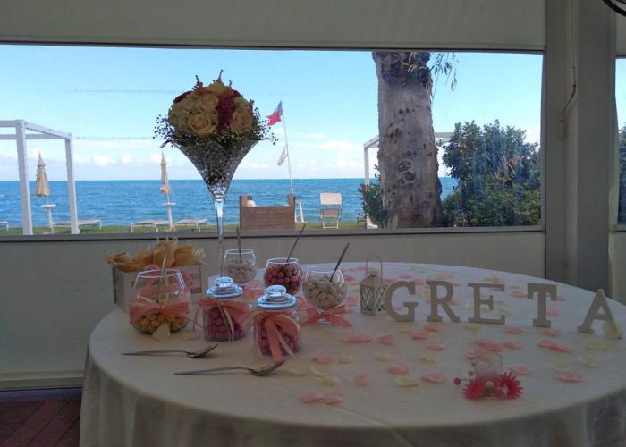 Tavolo decorato per festa con dolci e fiori, vista mare sullo sfondo.