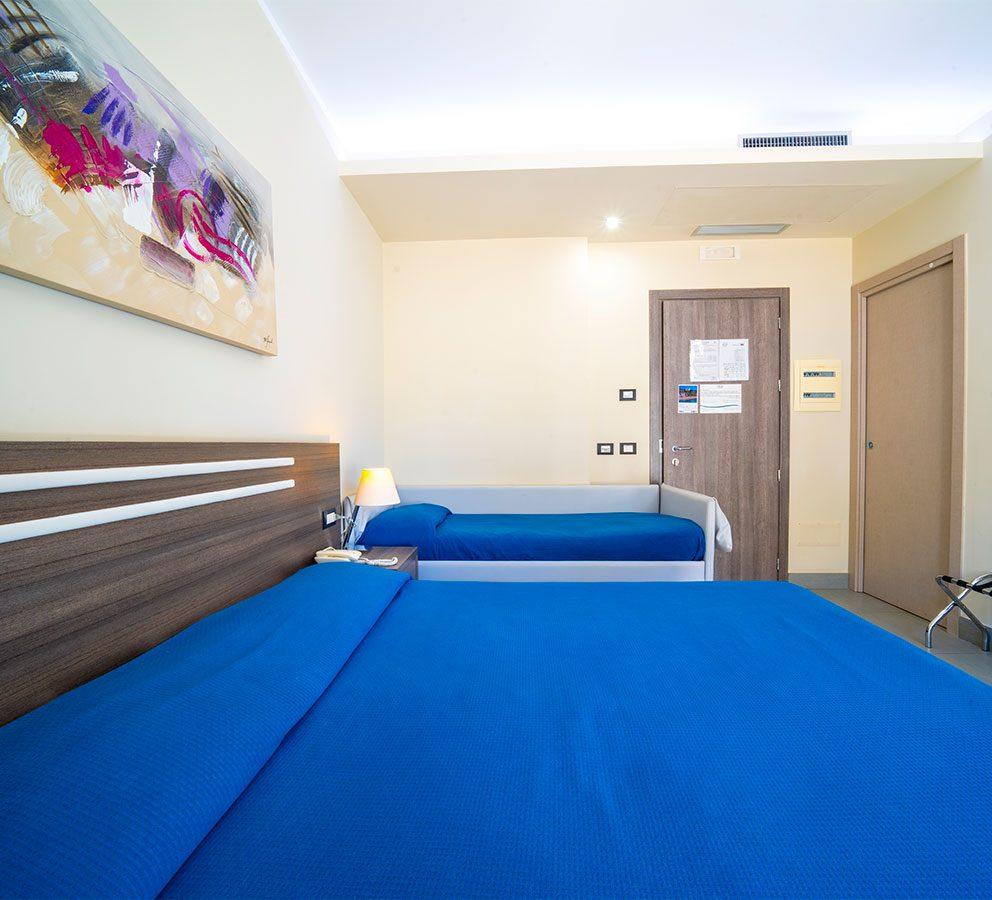 Camera d'albergo con letti blu, lampada e quadro astratto sulla parete.